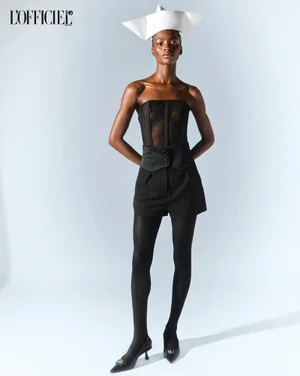 Model in black strapless corset top and shorts wearing a white paper hat against a light background