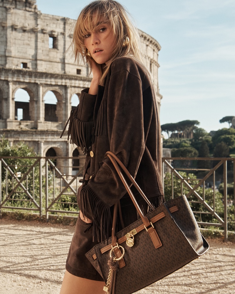 Model carrying a brown monogram tote bag and wearing a fringed suede jacket with the Colosseum in the background.