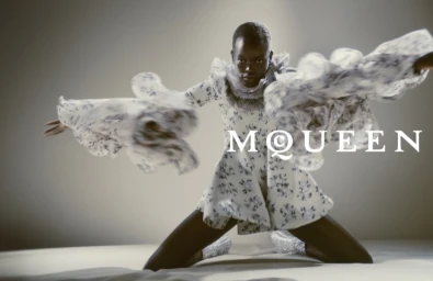 Model in a floral-print dress with ruffled sleeves kneeling against a neutral studio backdrop with branded text.