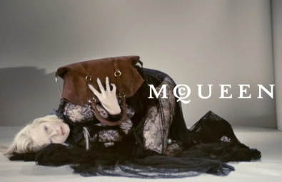 Model lying on studio floor in black lace dress holding a brown suede handbag for Alexander McQueen Fall-Winter 2025 campaign