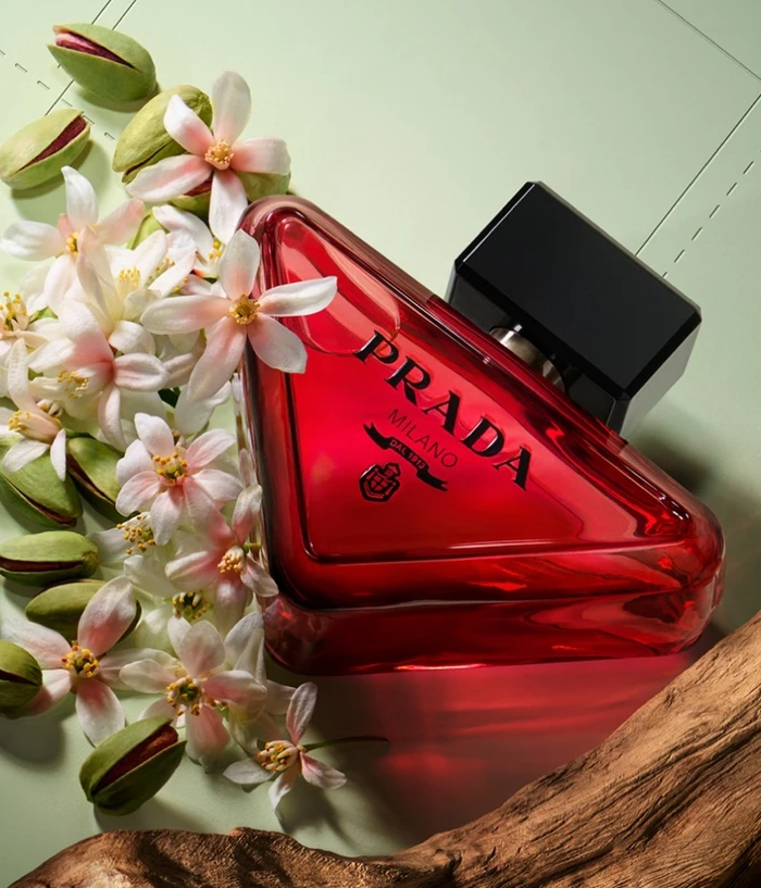 Prada perfume bottle with red liquid and black cap surrounded by white flowers and pistachio pods on a pale green surface.