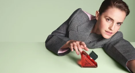 Emma Watson leaning over a surface reaching toward a triangular Prada perfume bottle against a pale green background