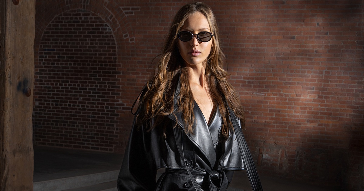 Angelina Kendall wearing black oval sunglasses and a belted black leather trench coat in front of an exposed red brick wall.