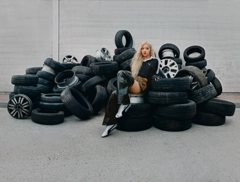 ROSÉ with blonde hair seated on a large pile of black car tires wearing a black top and silver Puma Speedcat sneakers.