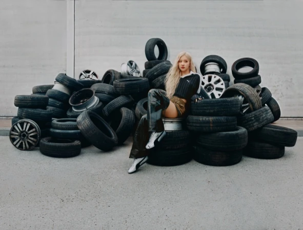 ROSÉ with blonde hair seated on a large pile of black car tires wearing a black top and silver Puma Speedcat sneakers.