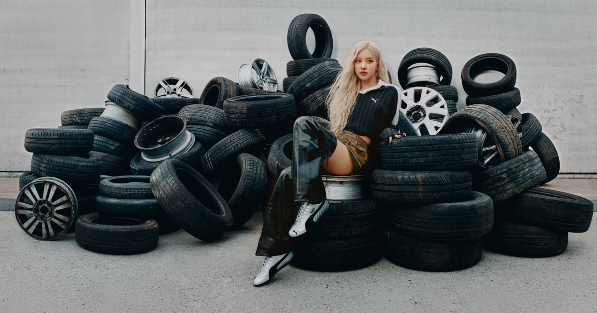 ROSÉ sits on a pile of stacked black car tires and silver rims wearing a cropped jacket and metallic Puma Speedcat sneakers.