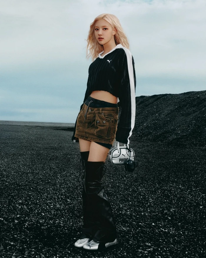 ROSÉ with blonde hair wearing a black crop top and brown cargo skirt standing against a dark gravel terrain and cloudy sky.