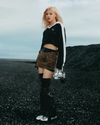 ROSÉ with blonde hair wearing a black crop top and brown cargo skirt standing against a dark gravel terrain and cloudy sky.