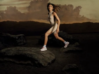 Zendaya running outdoors over dark rocks wearing a beige activewear romper and white sneakers against a cloudy sunset sky