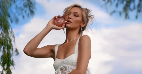 Bella Hadid holding a perfume bottle wearing a white lace top against a clear sky
