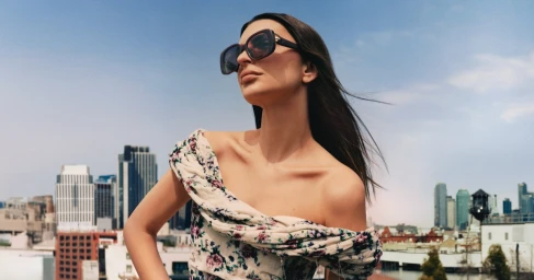 Emily Ratajkowski wearing oversized sunglasses and a floral off-shoulder dress against a city skyline backdrop for Tory Burch.