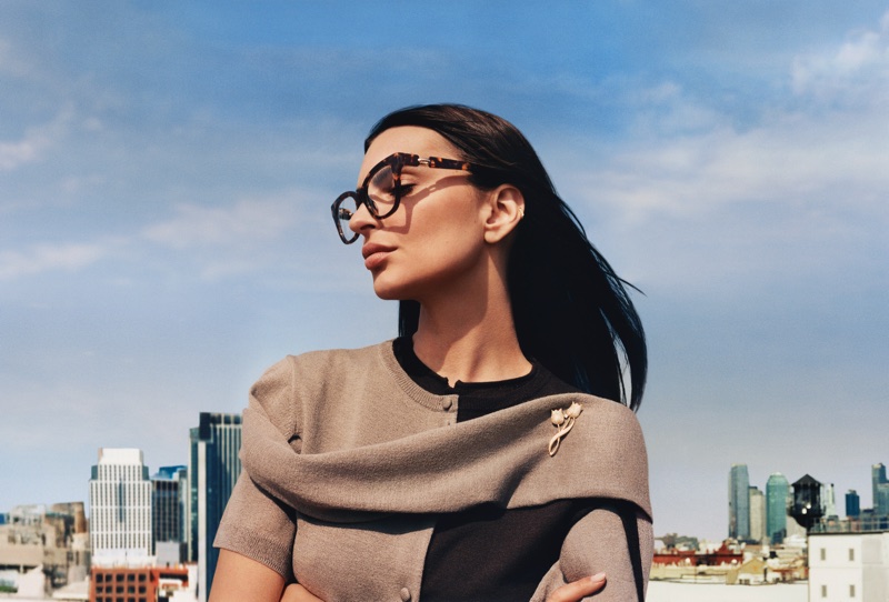 Emily Ratajkowski wearing tortoiseshell eyeglasses and a two-toned cardigan against an urban skyline backdrop