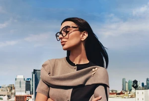 Emily Ratajkowski wearing tortoiseshell eyeglasses and a two-toned cardigan against an urban skyline backdrop