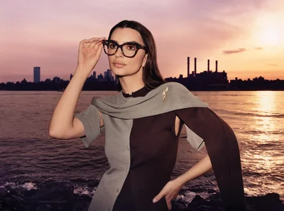 Emily Ratajkowski wearing oversized black frame glasses and a two-tone grey and brown top against an outdoor waterfront sunset backdrop.