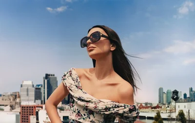Emily Ratajkowski wearing oversized sunglasses and a floral off-shoulder top against a city skyline backdrop.