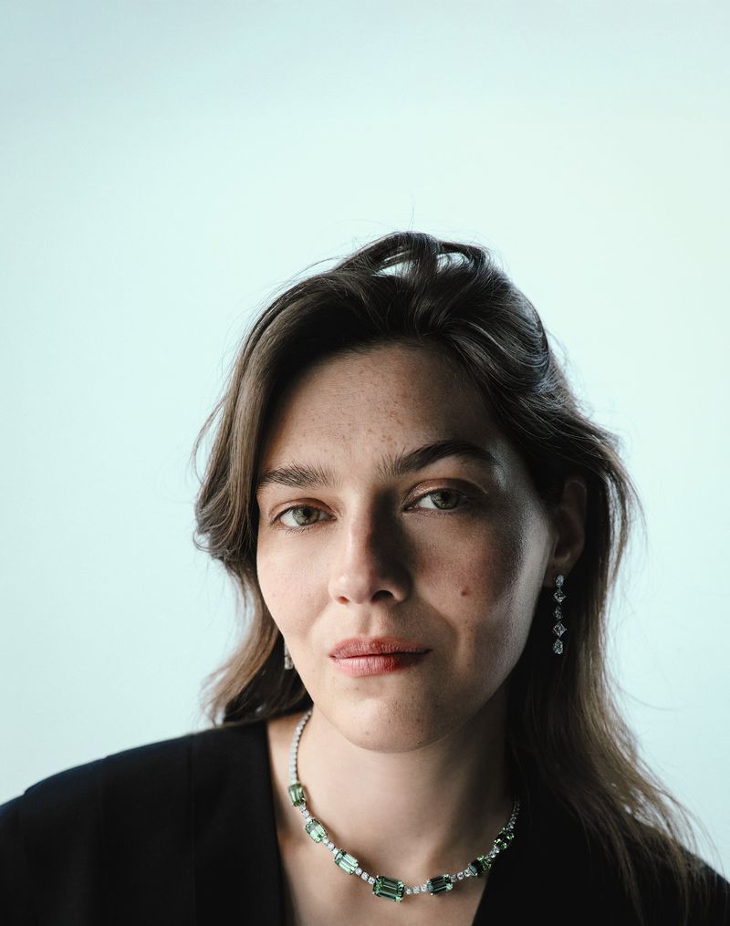 Close-up of a woman with green eyes and dark hair wearing a gemstone necklace and earrings against a light blue background.