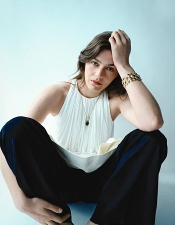 A young adult woman in a sleeveless white top and black pants poses in a studio with jewelry, seated against a light blue background.