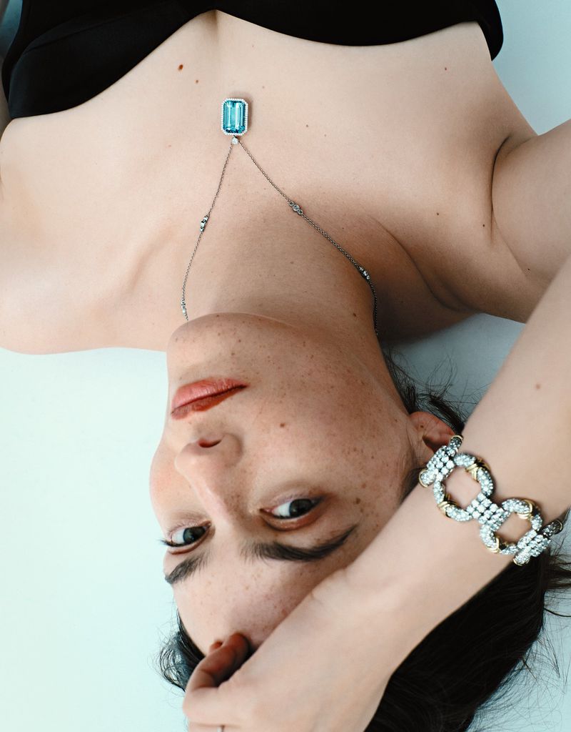 Adult female with freckles wearing a black top and jewelry, including a necklace and bracelet, posing against a light blue backdrop.