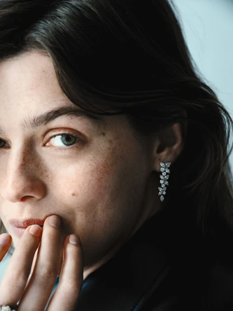 Close-up of a woman wearing flower-shaped diamond earrings, her fingers near her mouth, and a ring on her hand.