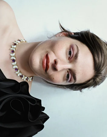 Close-up portrait of an adult woman with light skin, wearing a black strapless dress and an ornate jewel necklace in a light indoor setting.