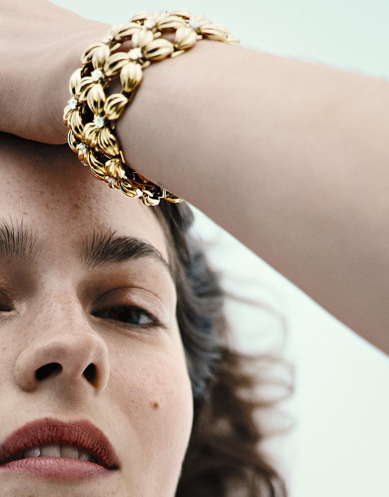 Close-up of a woman's face with a gold, floral-designed bracelet on her arm, set against a clean white background. Beauty and Jewelry.