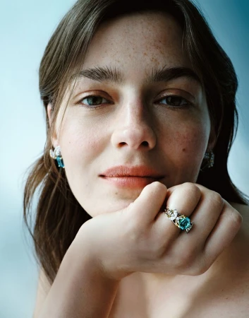 Close-up of a woman wearing turquoise jewelry including a ring and earrings, set against a muted blue background.