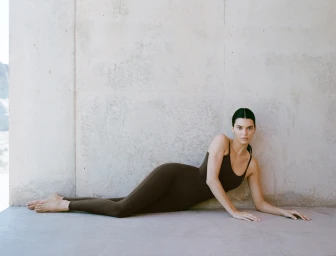 Model Kendall Jenner reclining on the ground wearing dark brown activewear against a plain concrete wall.