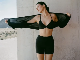 Kendall Jenner in a black sports bra and shorts holding a long-sleeved top against an architectural wall.