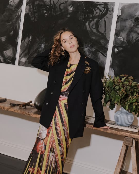 Cleo Wade wearing an oversized black blazer and patterned dress leaning against a wooden table in front of abstract artwork.