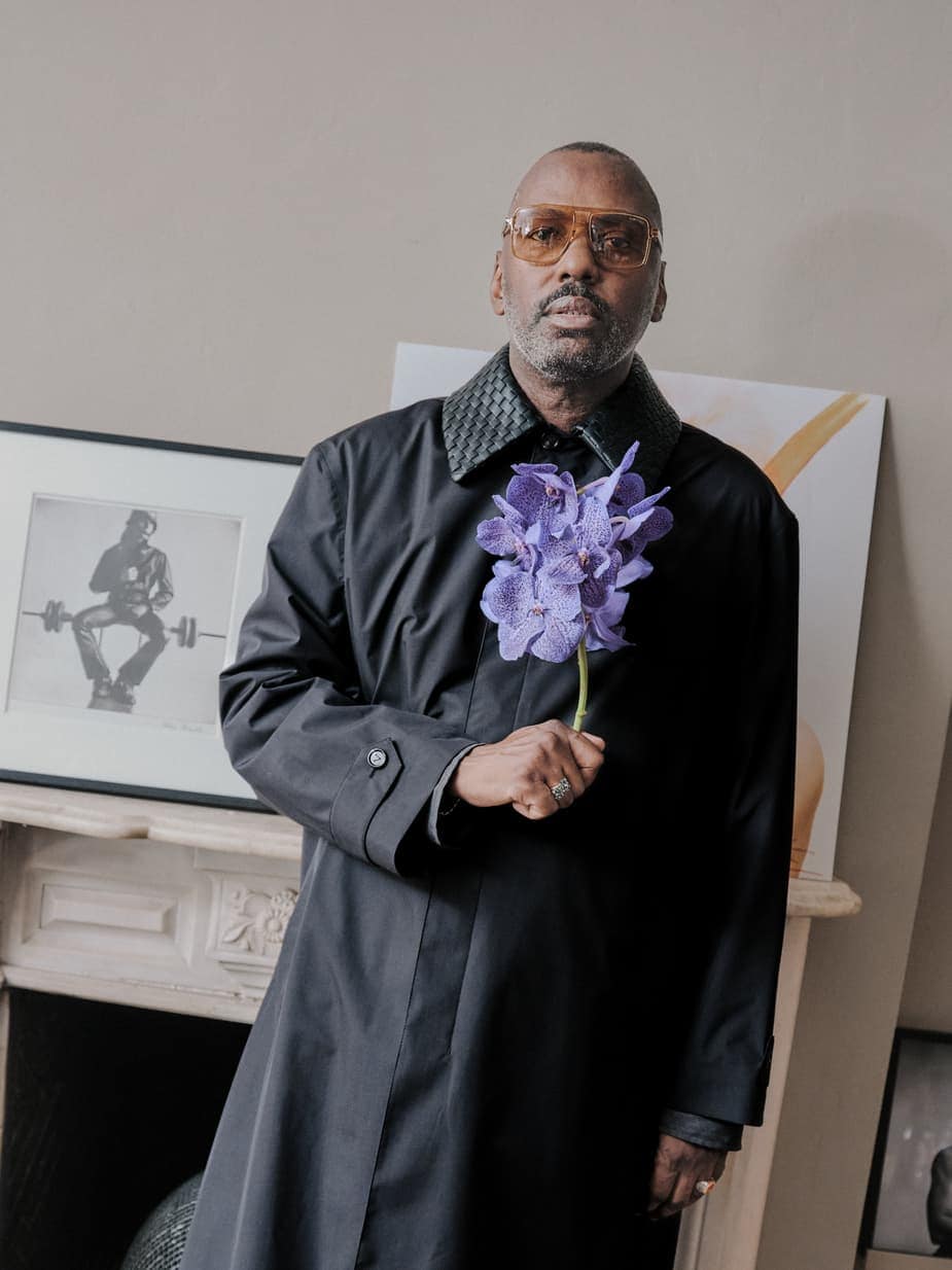 Stephen Galloway wearing tinted glasses and a black coat while holding a purple orchid stalk near a mantelpiece.