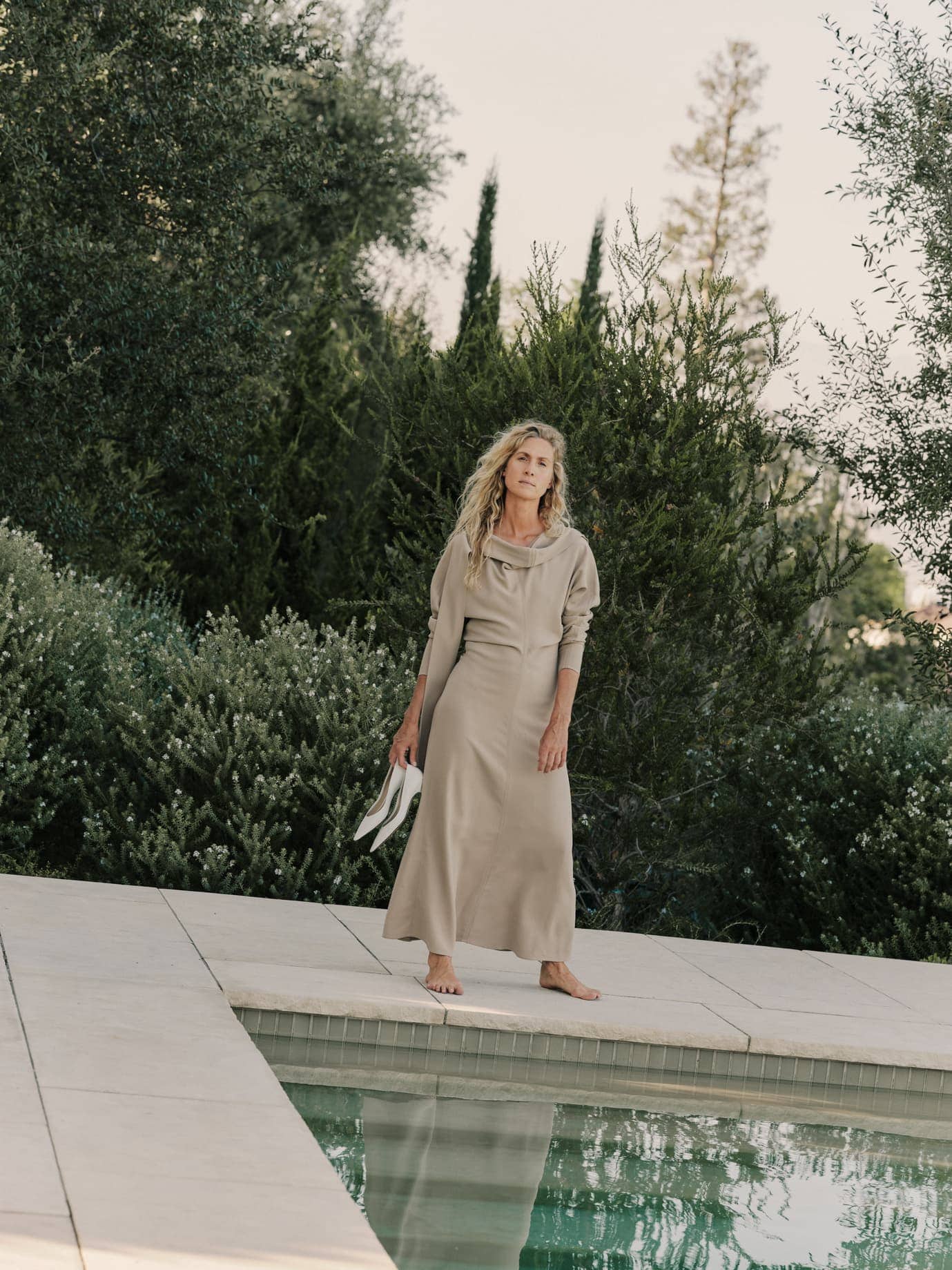 Woman in a long beige dress standing barefoot beside an outdoor pool holding a pair of white pointed heels.