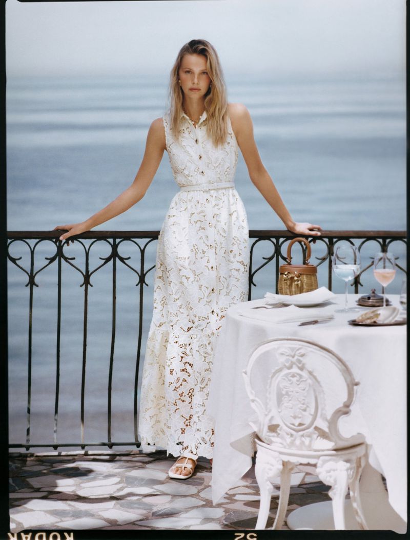 A fair-skinned adult woman in a white dress on a balcony overlooking the sea, alongside a table with glasses and plates. 