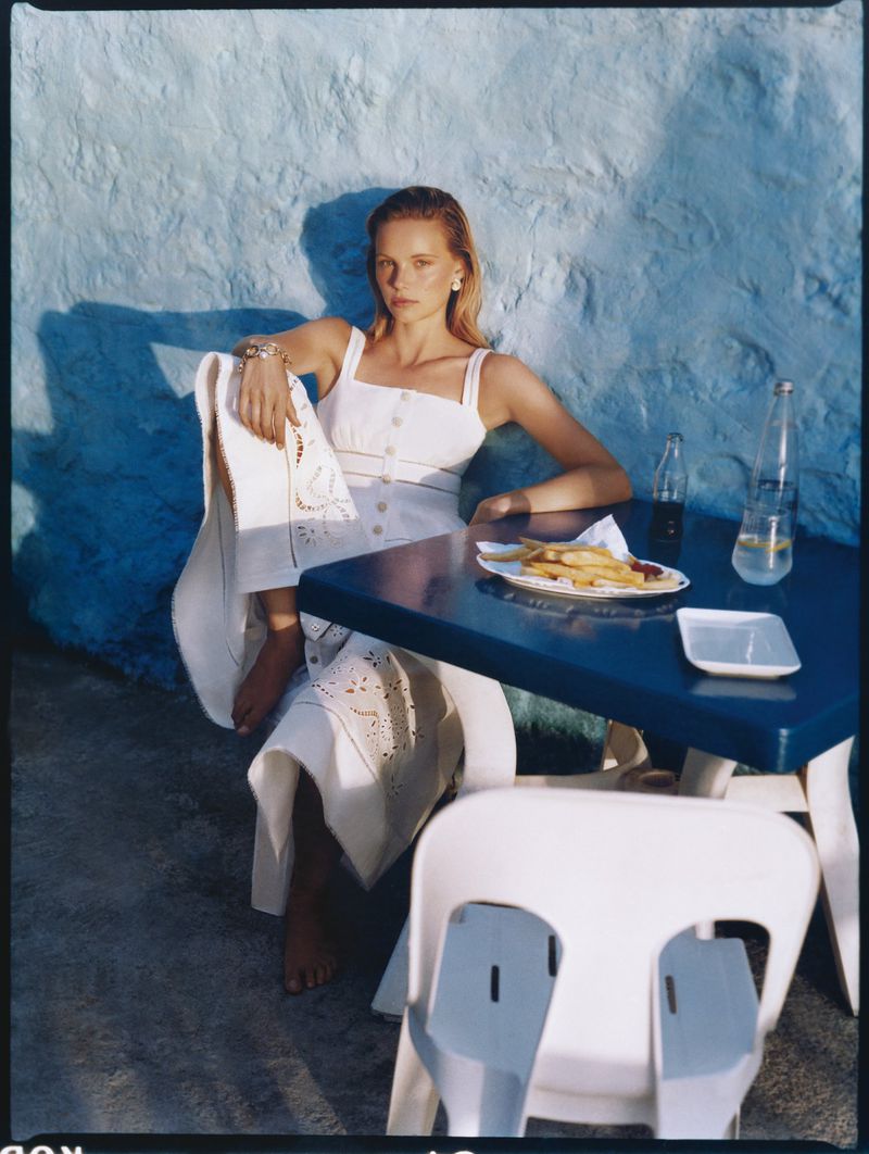 Adult woman in a white dress seated at a blue table outdoors. The scene includes drinks, fries, and a textured blue background wall.