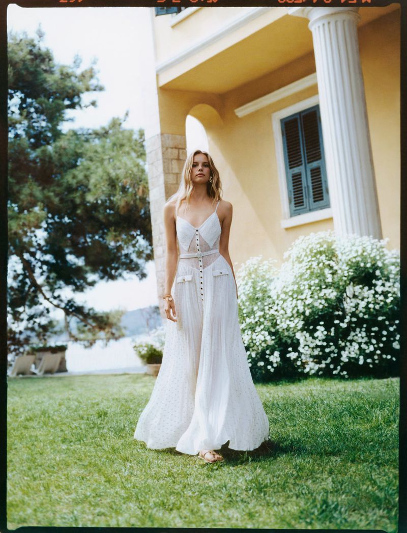 A blonde woman in a white dress walks across a green lawn toward a yellow building with white columns and green shutters.