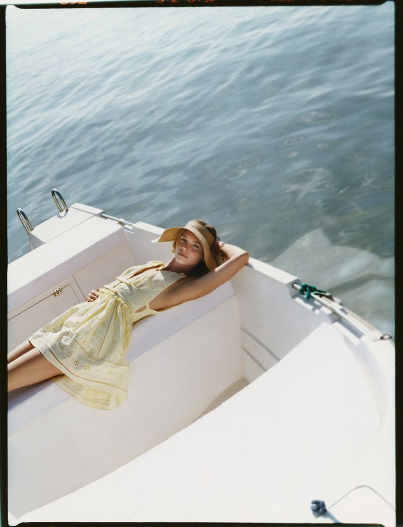Woman in yellow dress and sun hat reclines on a white boat, surrounded by rippling water on a bright, sunny day. Boat trip.