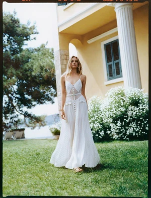 Model in a white sundress standing on a lush lawn in front of a yellow villa