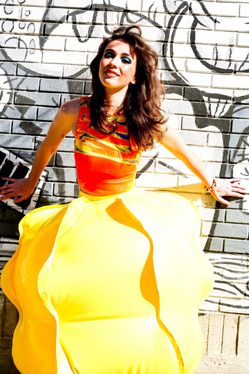 A cheerful woman in a vibrant orange top and large yellow skirt poses against a brick wall with graffiti art, bathed in sunlight.