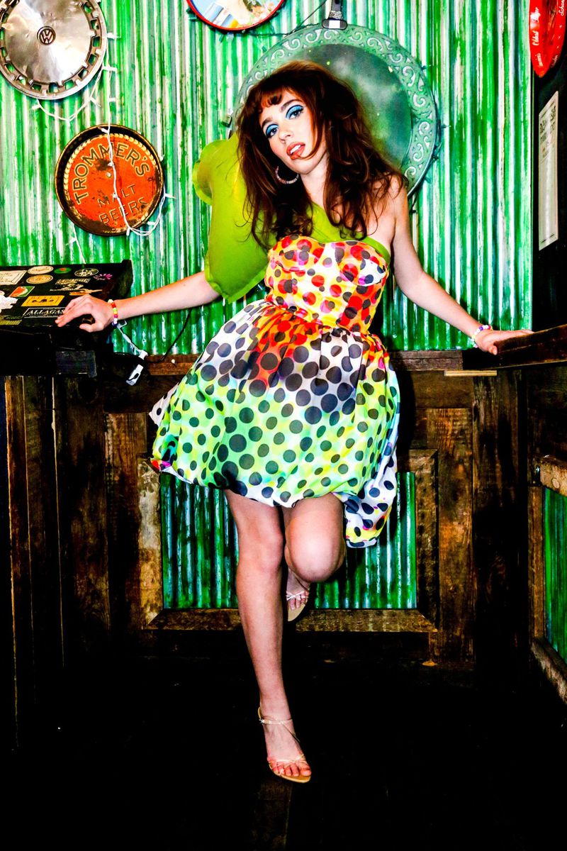 A young woman poses in a vibrant polka-dot dress, leaning against a rustic bar with a backdrop of corrugated metal and vintage decor.