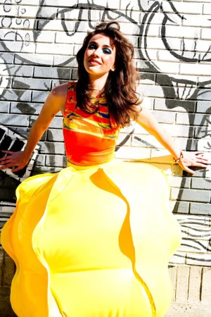 A woman posing in a voluminous yellow sculptural skirt and bright orange printed top against a graffiti-painted brick wall.
