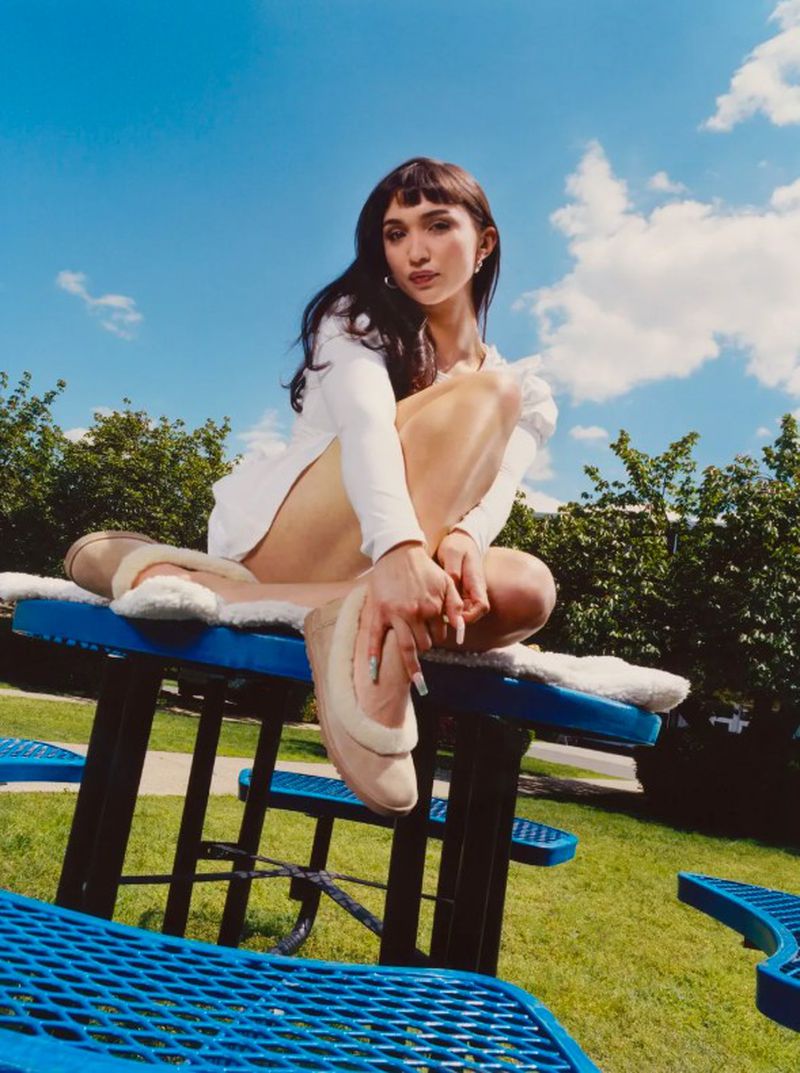 Rowan Blanchard seated on a blue picnic table wearing a white long-sleeve dress and beige shearling slip-on shoes under a blue sky.