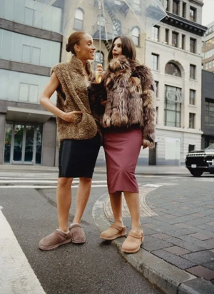 Adwoa Aboah and Emily Ratajkowski wearing textured outerwear and slip-on shoes on a city street under a clear umbrella.