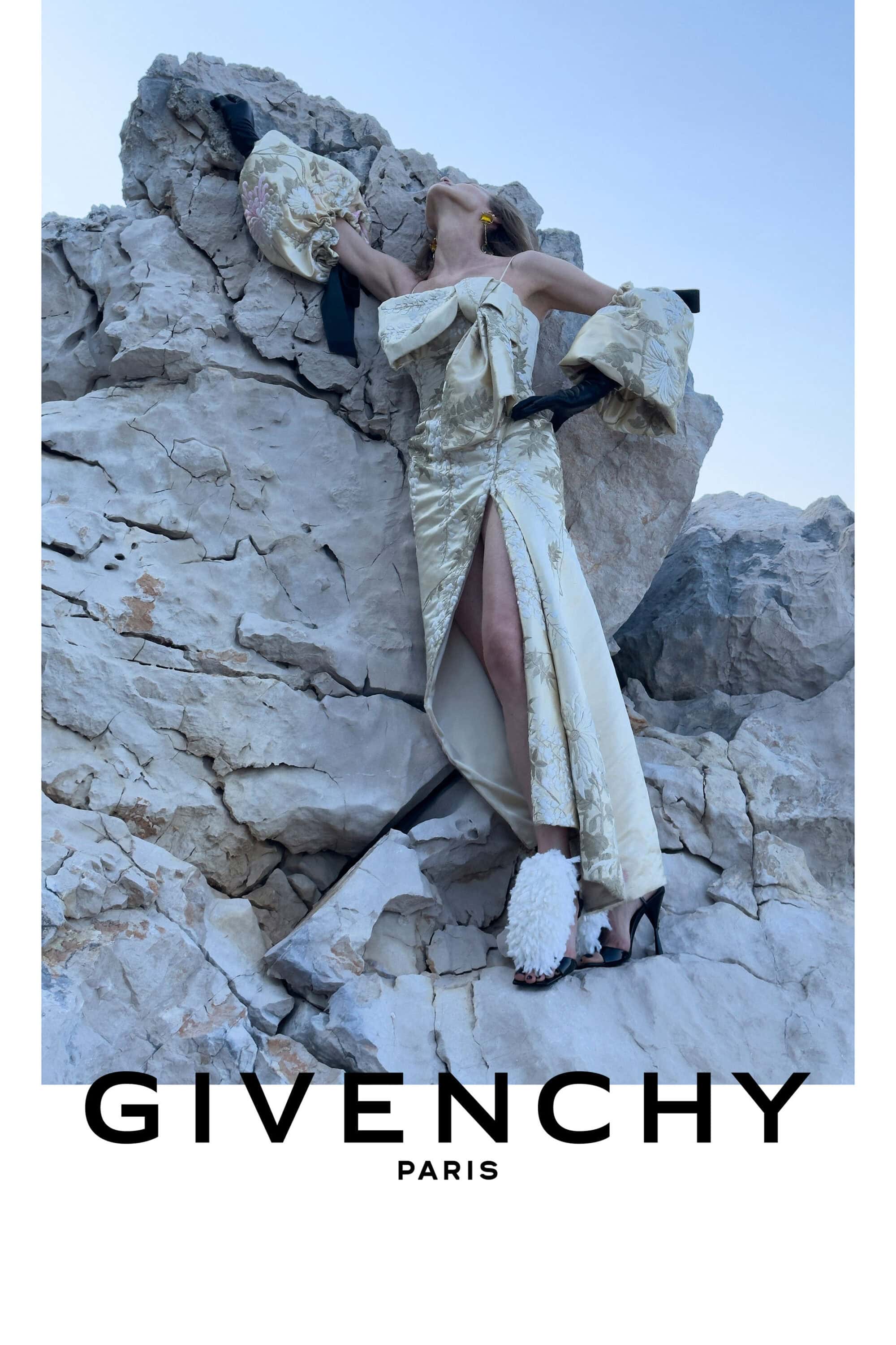 Model in a cream jacquard gown and black gloves leaning back against a pale rocky cliff face.