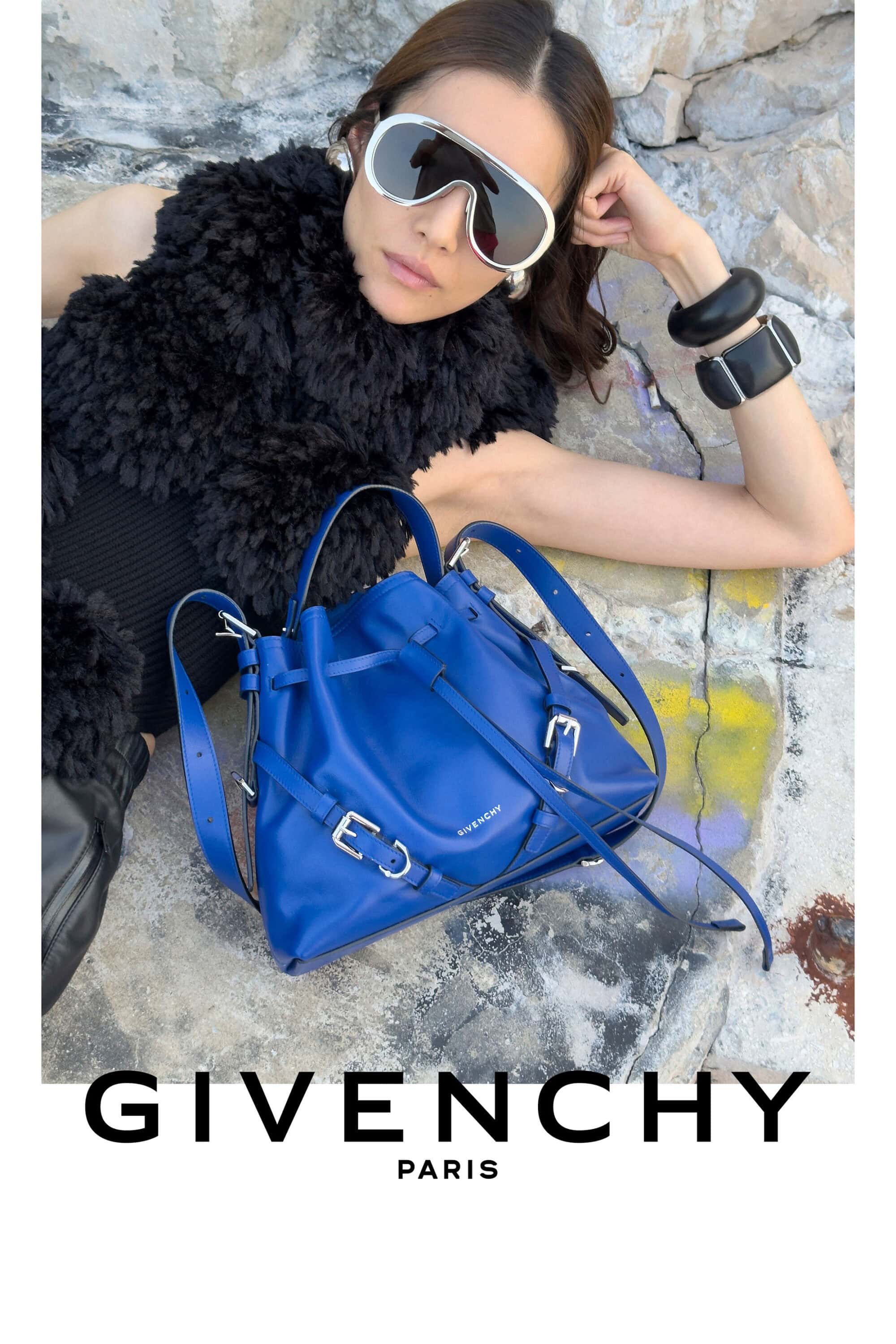 Model Liu Wen wearing shield sunglasses and a black textured top reclining beside a cobalt blue leather handbag on stone ground.
