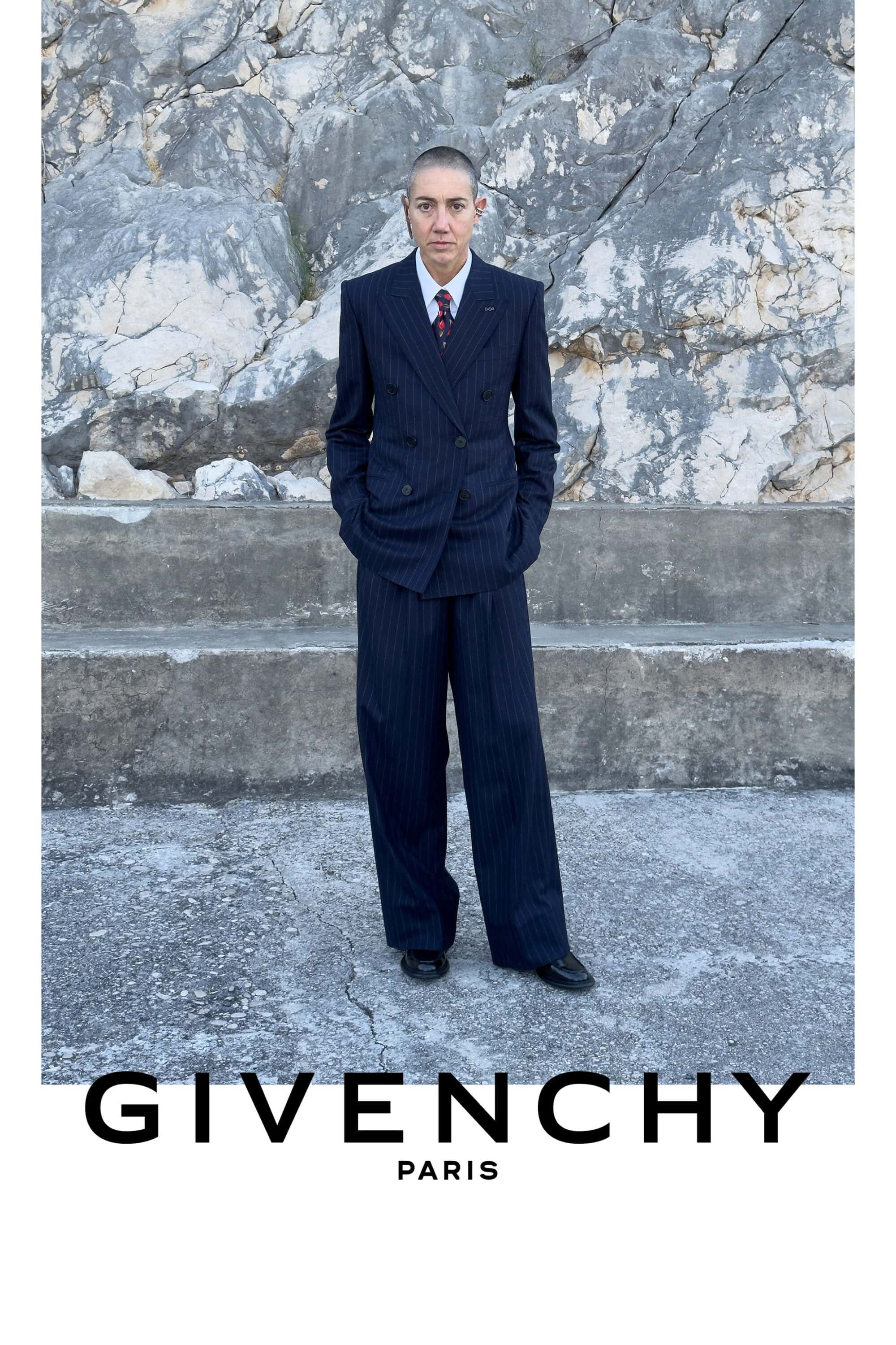 Constance Debré wearing a dark pinstripe double-breasted suit standing before a rocky wall for Givenchy Winter 2026.
