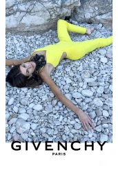 Model Kaia Gerber reclined on a pebble beach wearing a bright yellow jumpsuit and black sculptural chain necklace for Givenchy.