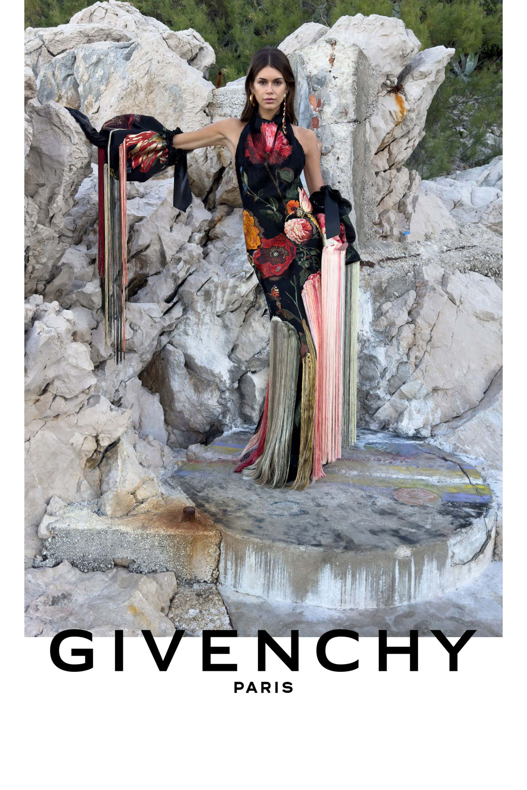 Kaia Gerber wearing a black floral halter gown with long fringe accents against pale rocky cliffs for Givenchy.