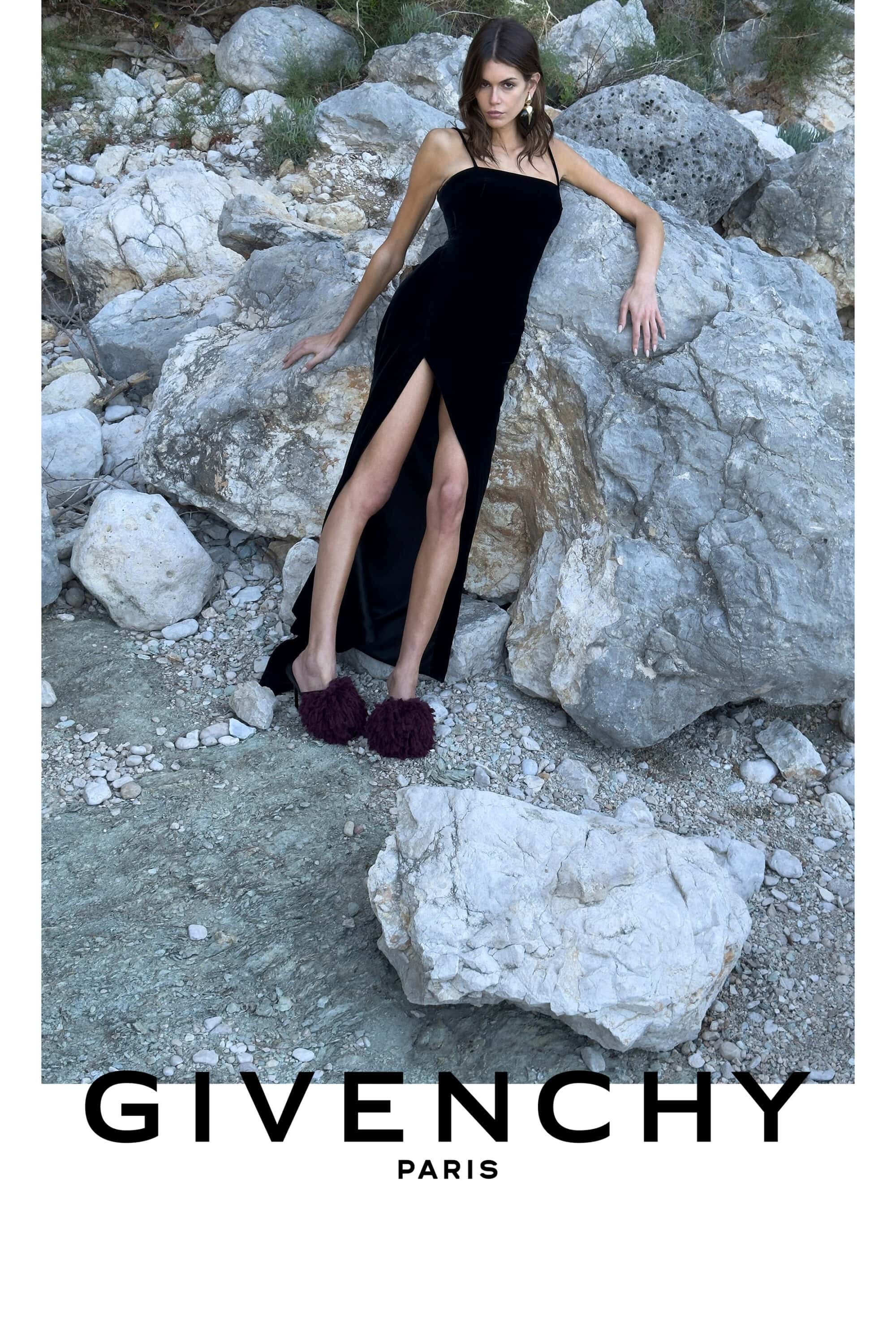 Kaia Gerber wearing a black slit dress and dark fuzzy slippers leaning against large rocky boulders for Givenchy.