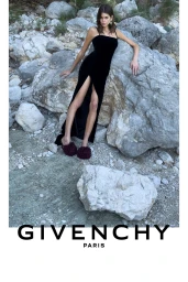 Kaia Gerber wearing a black slit dress and dark fuzzy slippers leaning against large rocky boulders for Givenchy.