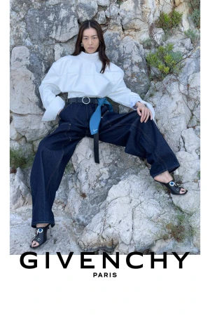 Liu Wen seated against a rocky cliff wearing an oversized white blouse and wide-leg dark denim trousers for Givenchy.