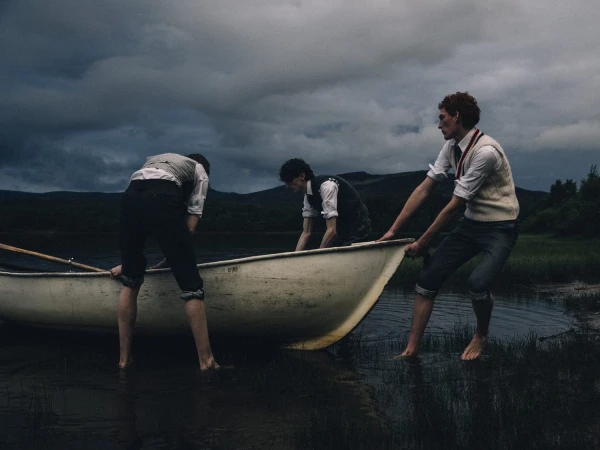 Three men in tailored knitwear and rolled trousers pulling a white rowboat ashore in dark lake water under overcast skies.